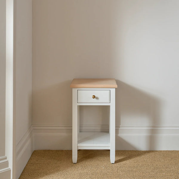 A small white nightstand with a light natural wood top, a single drawer with a round brass knob, and an open lower shelf, set against a neutral beige wall on a woven jute rug.