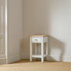 A small white side table with a natural wood top and a single drawer, set in the corner of a room with beige walls and a textured beige carpet.