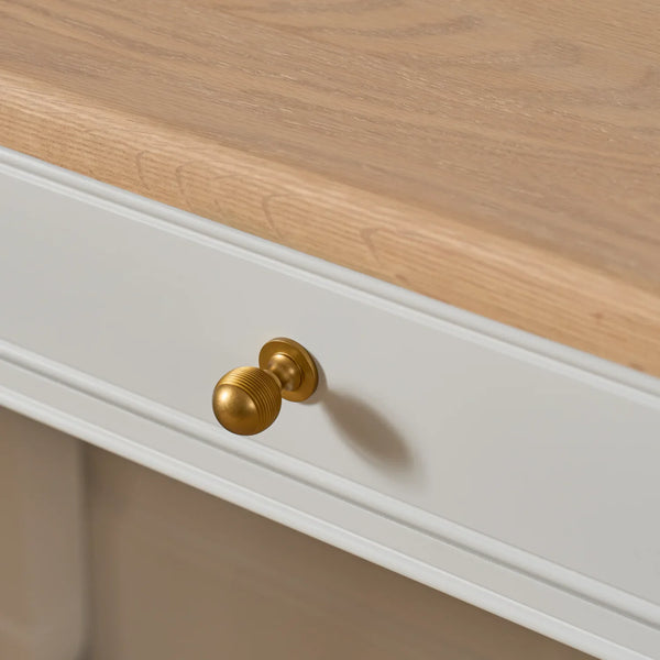 A close-up of a white drawer front with a round brushed brass knob, set beneath a light natural wood tabletop.