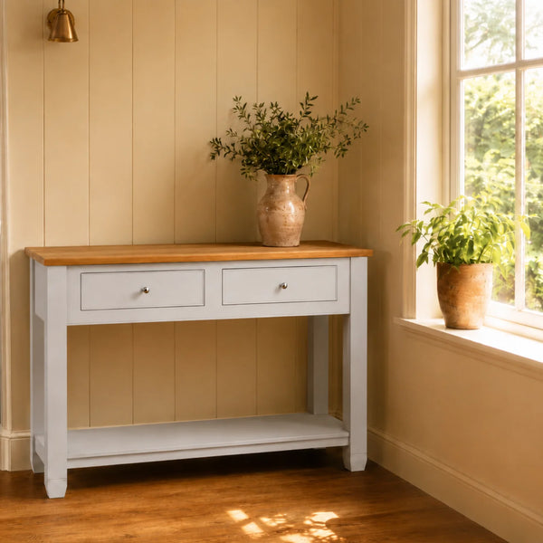 A pale grey console table with a natural oak top, styled in a warm, sunlit room with light beige panelled walls, a window, and potted plants.