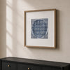 Framed wall art hanging on a beige wall above a dark wood console table. The artwork shows a circular blue patterned print with a white mat and a light wood frame.