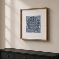 Framed wall art hanging on a beige wall above a dark wood console table. The artwork shows a circular blue patterned print with a white mat and a light wood frame.