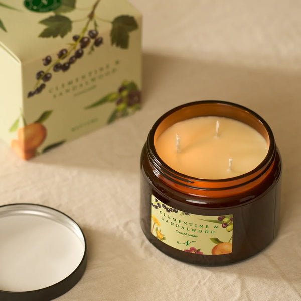 An amber glass jar candle with a cream-colored wax fill and three wicks, shown open with its lid beside it, next to its illustrated product box on a beige fabric surface.