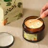 An amber glass jar candle with three lit wicks and a cream-colored label reading Clementine & Sandalwood, shown on a beige fabric surface. A hand is lighting the candle with a wooden match, and the matching product box is blurred in the background.