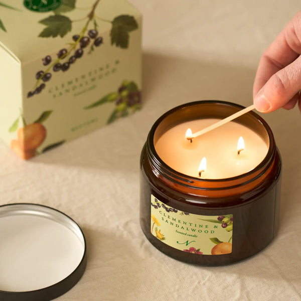 An amber glass jar candle with three lit wicks and a cream-colored label reading Clementine & Sandalwood, shown on a beige fabric surface. A hand is lighting the candle with a wooden match, and the matching product box is blurred in the background.