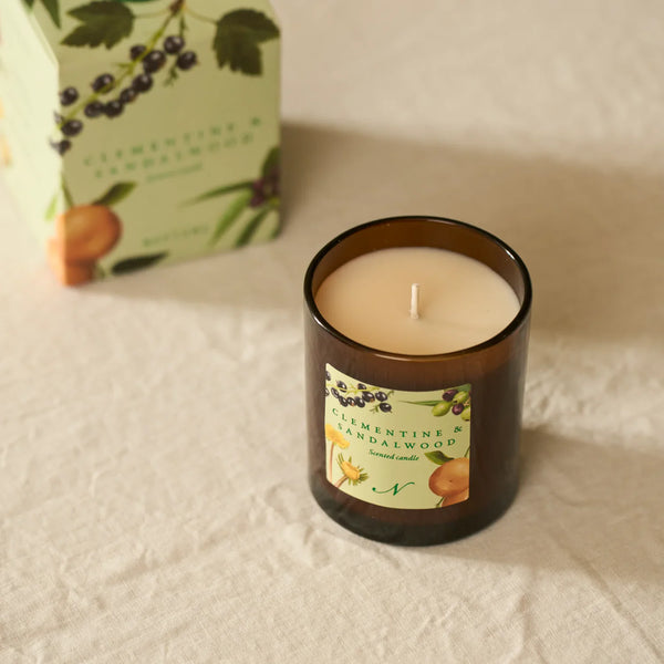 An amber glass jar candle with cream-colored wax sits on a textured white fabric surface, with its illustrated product box blurred in the background. The label reads Clementine & Sandalwood.