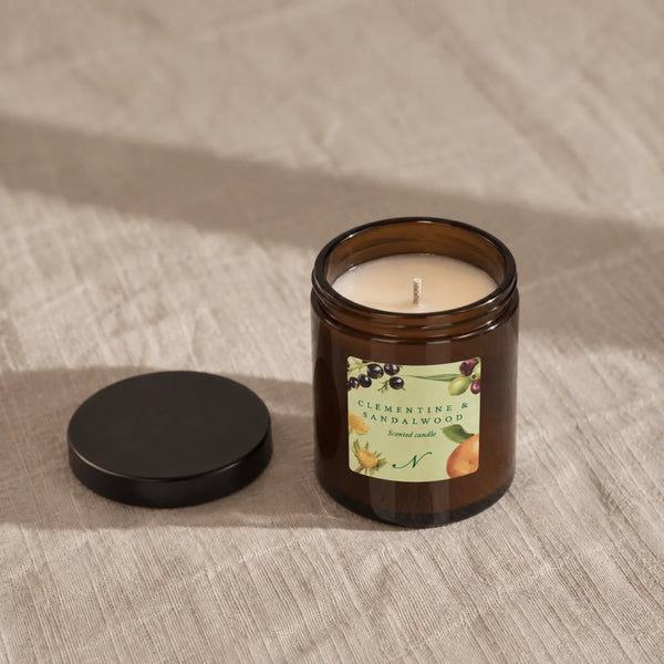 An amber glass jar candle with cream-colored wax sits on a textured white fabric surface. The label reads Clementine & Sandalwood.