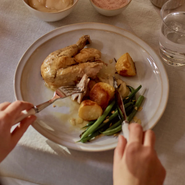 A plated meal on a light beige ceramic plate, featuring roasted chicken, roasted potatoes, green beans, and a drizzle of sauce, with hands holding a fork and knife at the bottom of the frame.