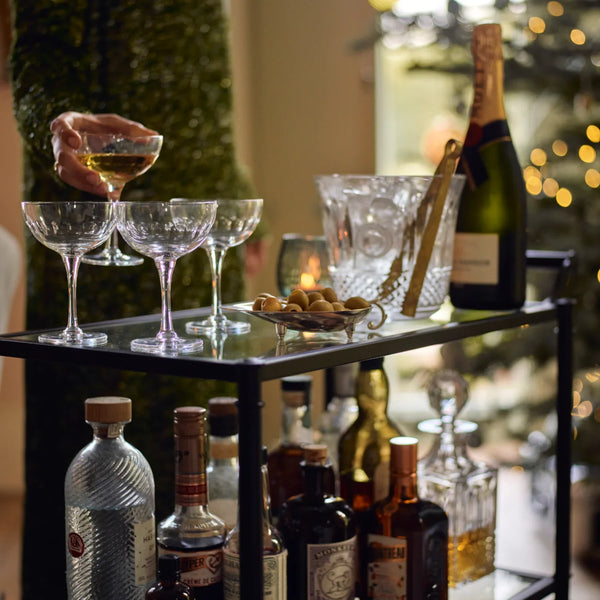 A black metal bar cart styled with an assortment of bottles, crystal glasses, a champagne bucket, and olives, with a hand holding a wine glass in the foreground against a festive holiday background.