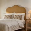 A styled bedroom scene featuring a bed with a tall, arched upholstered headboard in a warm tan or camel color, dressed in white bedding with decorative patterned pillows and white scalloped trim. A matching wooden nightstand with a lit table lamp sits on the right against a warm beige wall.