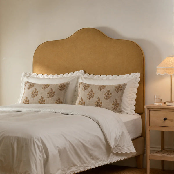 A styled bedroom scene featuring a bed with a tall, arched upholstered headboard in a warm tan or camel color, dressed in white bedding with decorative patterned pillows and white scalloped trim. A matching wooden nightstand with a lit table lamp sits on the right against a warm beige wall.