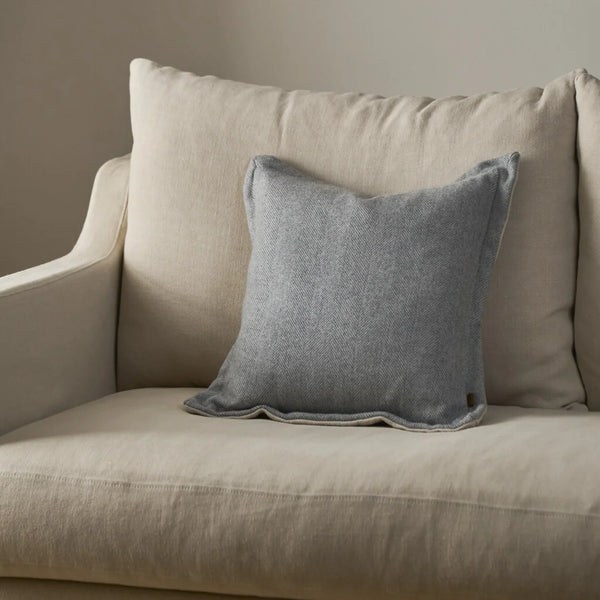 A square gray throw pillow with a textured linen-like fabric, styled on a beige upholstered armchair against a neutral beige wall.