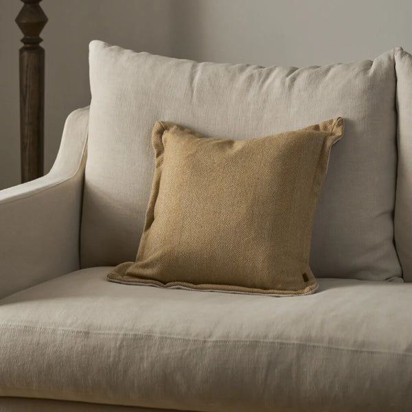 A square tan linen throw pillow with ruffled edges resting on a light beige upholstered armchair.