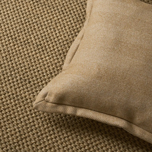 A close-up of a beige woven throw pillow with a subtle herringbone pattern, resting on a textured brown woven surface.