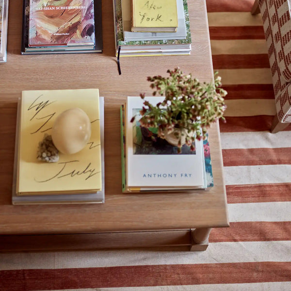A styled top-down view of a wooden coffee table holding several books, including a yellow 'My 1st July' book, a white 'Anthony Fry' book, and a small potted plant, with a striped rug underneath.