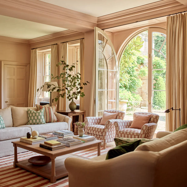 A beautifully styled living room featuring a light beige sofa, two patterned armchairs, a wooden coffee table, and large arched windows looking out onto a lush green garden. The room is decorated in warm beige and cream tones with striped curtains and a striped rug.