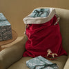 A red velvet Christmas gift sack with a beige drawstring opening sits on an olive green armchair, with wrapped presents and a small rocking horse nearby in a warm indoor setting.