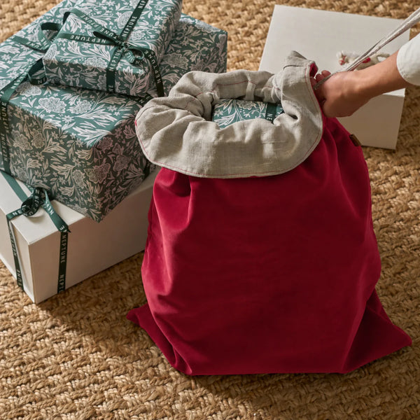 A red fabric gift bag with a light gray drawstring opening, being held open by a person's hand, with wrapped presents and white boxes on a woven jute rug.