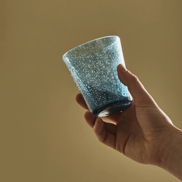 A single small blue textured glass tumbler with a speckled finish, held in one hand, shown with a netural background.