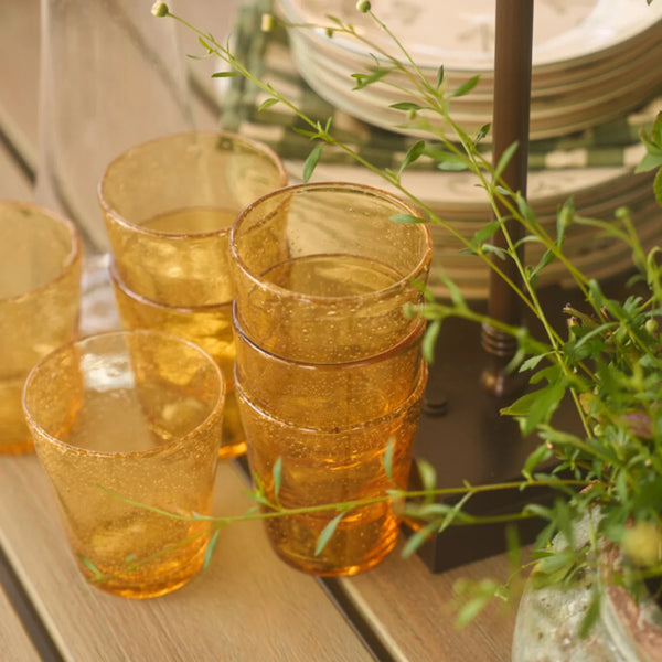 A set of amber-colored, textured glass tumblers arranged on a wooden table with green foliage in the foreground and stacked plates in the background.