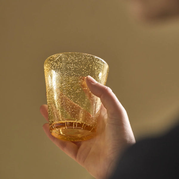 A hand holding a small, clear glass tumbler filled with shimmering gold glitter against a warm, solid beige background.