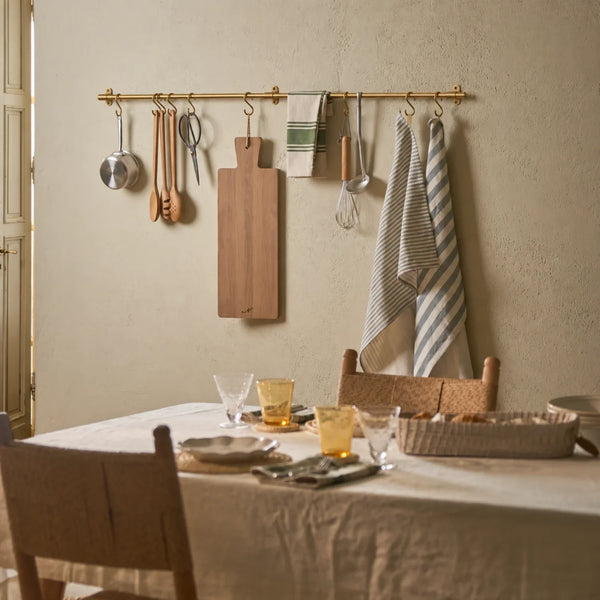 A brass wall-mounted hanging rack in a warm, rustic dining room setting, holding kitchen utensils, a cutting board, and striped towels above a set dining table.