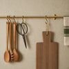 A brass wall-mounted hanging rack, holding kitchen utensils, a cutting board, and striped towels.