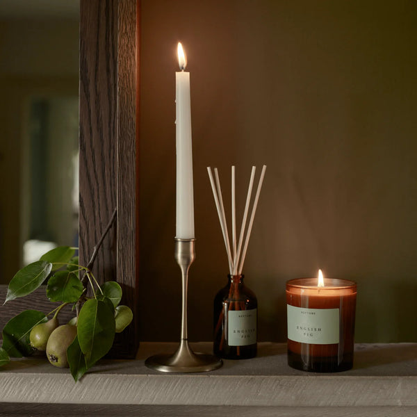 A styled interior scene on a shelf with two lit candles, a reed diffuser bottle, a tall white taper candle in a brass holder, and a small amber bottle with reeds. A branch with green leaves and small fruit rests on the left against a dark brown wall.