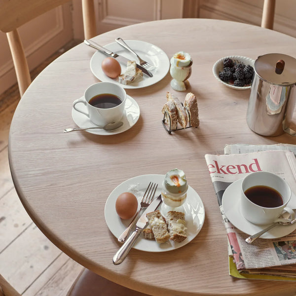 A round wooden dining table set with two white cups of black coffee on saucers, plates with an egg and toast, a small dessert, a metal pitcher, and a folded newspaper on a plate setting.