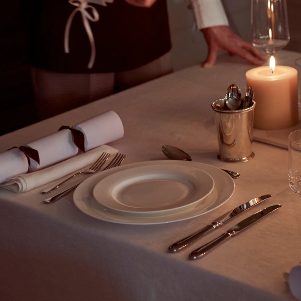 A dimly lit dining table set with white plates, silver cutlery, a rolled napkin with a dark ribbon, a silver spoon holder, and a lit pillar candle. A person in a dark apron is partially visible in the background.