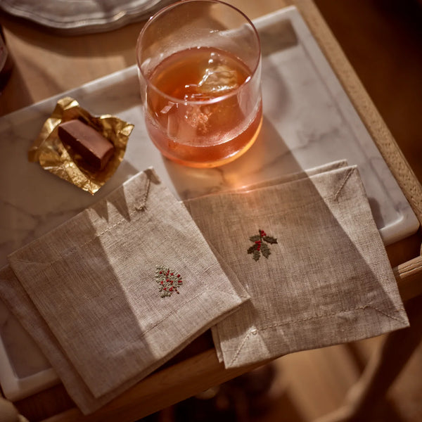 Two beige linen napkins with delicate embroidered holly motifs, arranged on a marble tray next to a glass of amber drink and a wrapped chocolate.