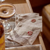 Two beige linen napkins with red embroidered monogram designs, styled on a wooden tray next to a cocktail glass.