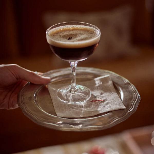 A hand holds a clear glass pedestal tray carrying a stemmed coupe glass filled with a layered dark beverage topped with a thick cream-colored foam head, set against a warm, softly blurred indoor background.