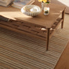 A light oak wood coffee table with a woven lower shelf, styled with a white seashell bowl, books, and a candle on a striped beige and brown rug.