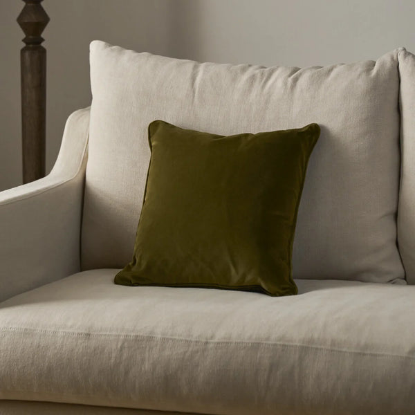 An olive green velvet square throw pillow resting on a light beige upholstered armchair.