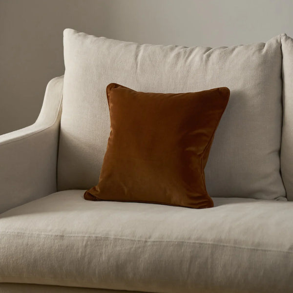 A square rust-brown velvet throw pillow resting on a light beige upholstered armchair against a neutral gray wall.