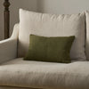 An olive green rectangular throw pillow resting on a light beige upholstered armchair in a softly lit indoor setting.