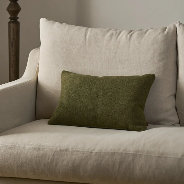 An olive green rectangular throw pillow resting on a light beige upholstered armchair in a softly lit indoor setting.