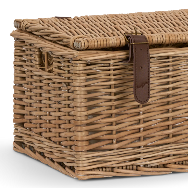 A close-up of a natural wicker storage chest with a hinged lid, featuring a dark brown leather strap and brass buckle on the front.