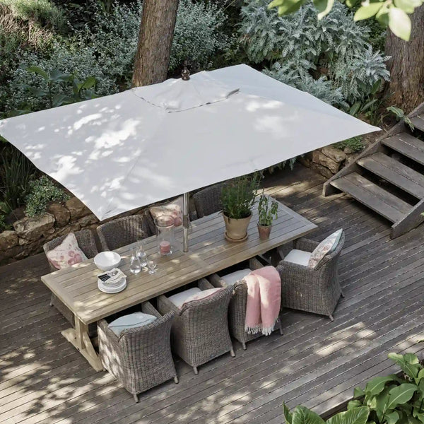 Outdoor patio dining set with a large square white patio umbrella, a wooden dining table, and four woven gray armchairs on a wooden deck surrounded by green plants.
