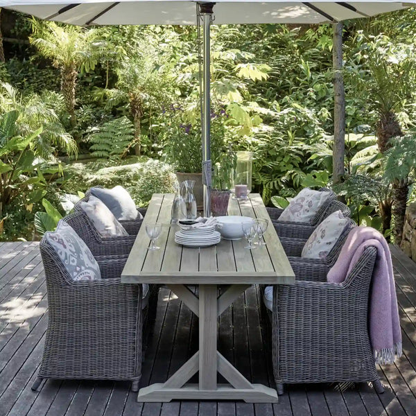 Outdoor dining set on a wooden deck surrounded by lush greenery, featuring a gray wicker dining table with six matching armchairs, a large white patio umbrella, and tableware set for four.