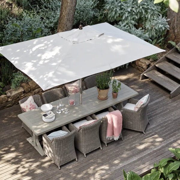 Outdoor patio dining set with a rectangular light gray table, woven gray chairs with pink patterned cushions, a large off-white patio umbrella, and a pink throw blanket on a wooden deck surrounded by greenery.