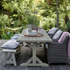 An outdoor dining set on a wooden deck surrounded by lush green garden foliage. The set includes a light gray rectangular slat-top table with a trestle base, a gray woven armchair with a pink throw blanket, a matching bench with a patterned cushion, and a side table with patterned cushions.
