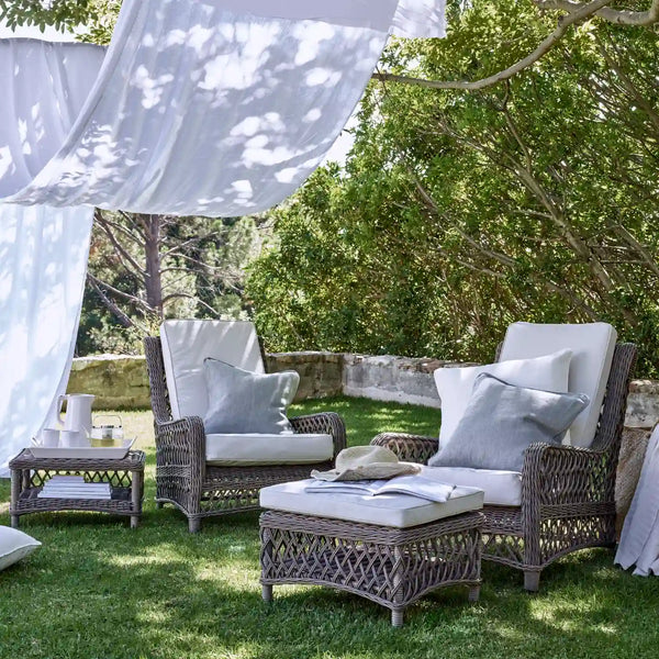 Outdoor patio furniture set on a grassy lawn under draped white fabric, featuring a wicker armchair, matching ottoman, side table, and cushioned lounge chair with white and gray pillows, surrounded by trees.