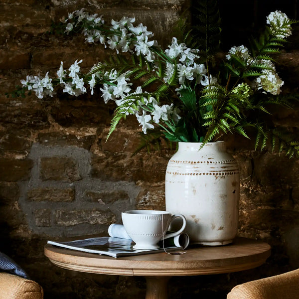 A rustic ceramic vase filled with white flowers and green ferns sits on a round wooden table beside a white teacup and a magazine, set against a dark stone wall.