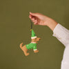 A hand wearing a white long-sleeve shirt holds up a small felt dog Christmas ornament dressed in a green sweater and hat against an olive green background.