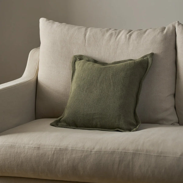 An olive green textured throw pillow resting on a light beige upholstered armchair against a neutral gray wall.