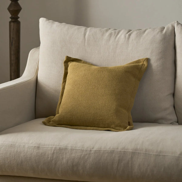 A square saffron yellow throw pillow with a textured linen-like fabric, styled on a light beige upholstered armchair.