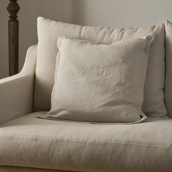 A light beige textured throw pillow resting on a matching beige upholstered armchair in a softly lit interior setting.