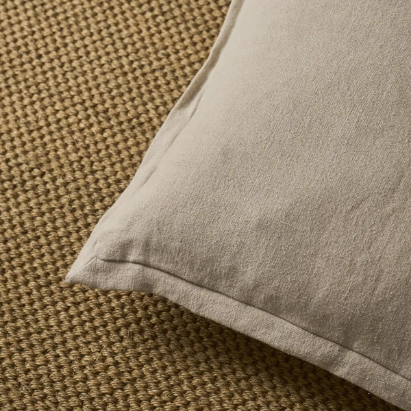 A close-up of a light beige pillow resting on a textured brown woven rug.